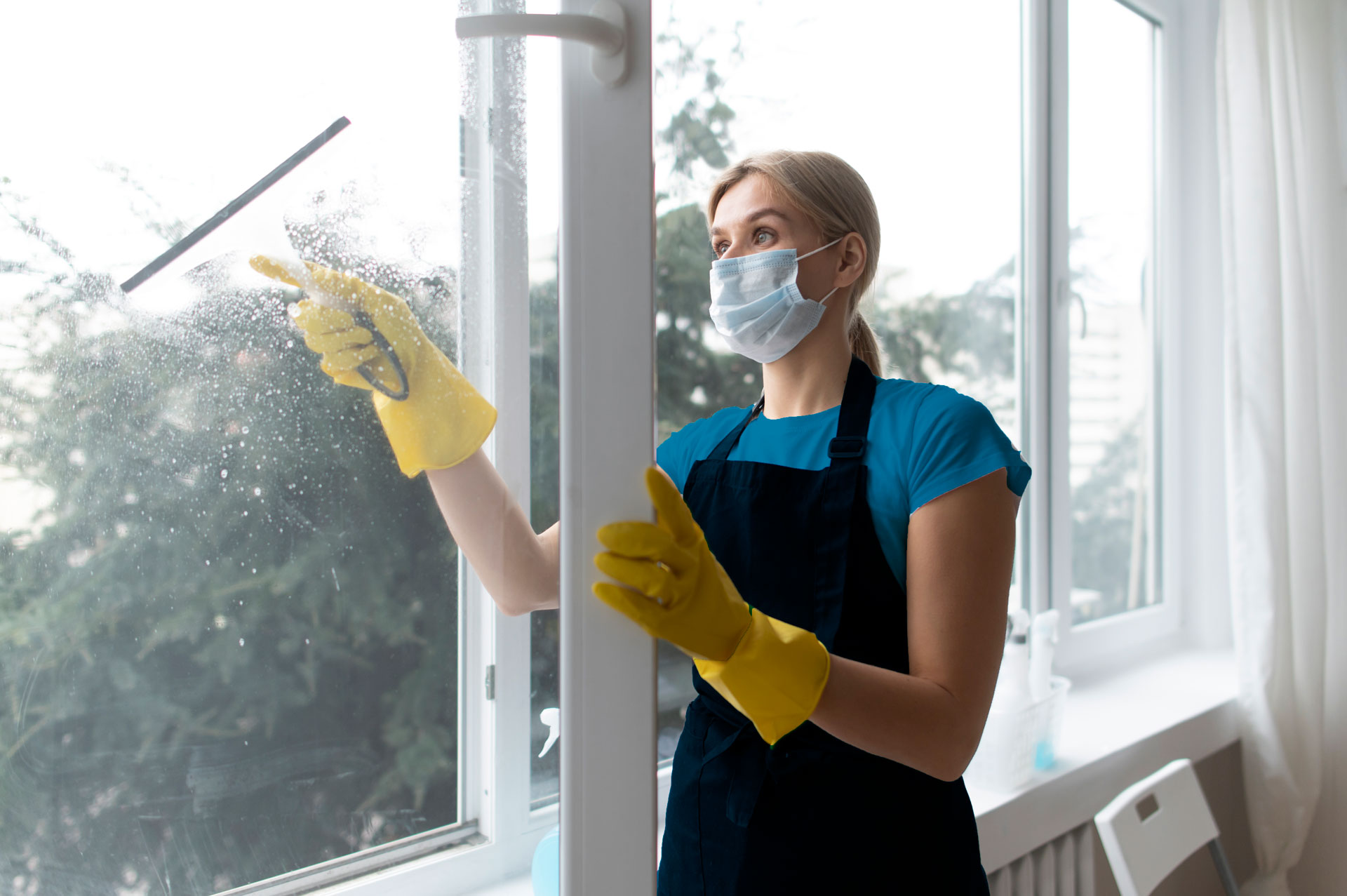 A maid from a cleaning service in Austin Texas wearing a mask and gloves cleaning a window.